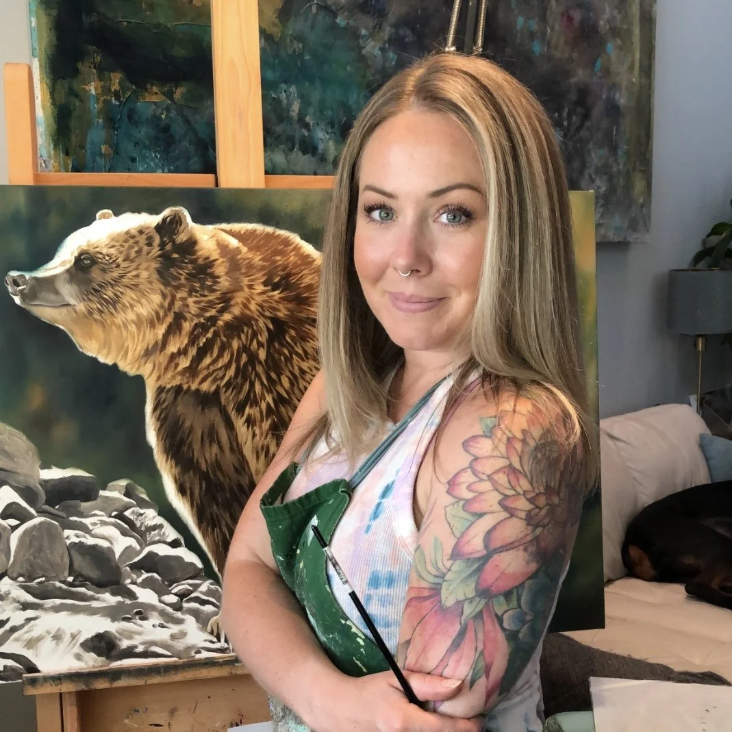 Erin Burge in her studio beside an oil painting of a grizzly bear, holding a paintbrush